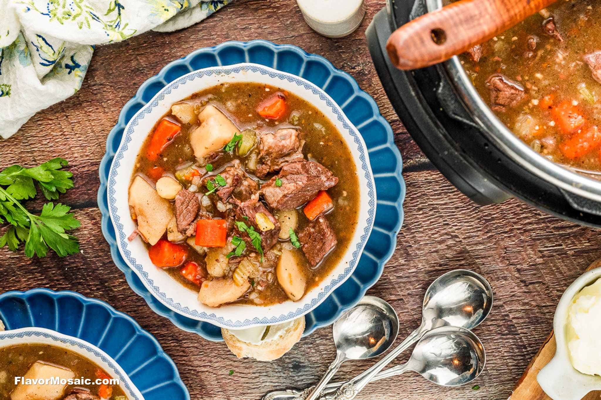 A bowl of Instant Pot Beef Stew with chunks of beef, potatoes, and carrots, garnished with herbs. The bowl is on a wooden table beside parsley, spoons, and another pot of stew.
