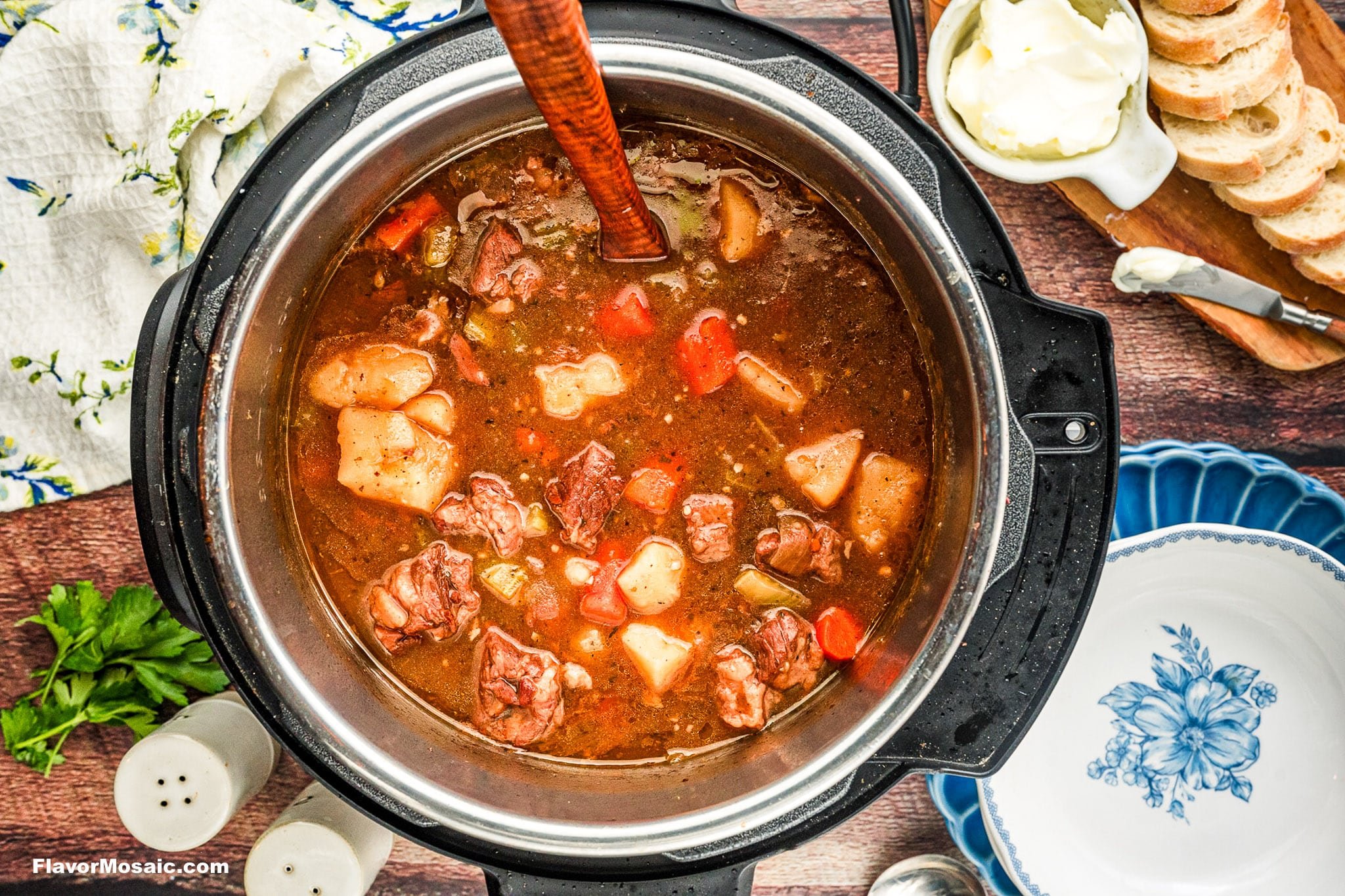 A top-down view of a pot of hearty Instant Pot Beef Stew with chunks of beef, potatoes, and carrots, next to a bowl, sliced bread, a dish of butter, salt and pepper shakers, and parsley on a wooden table.