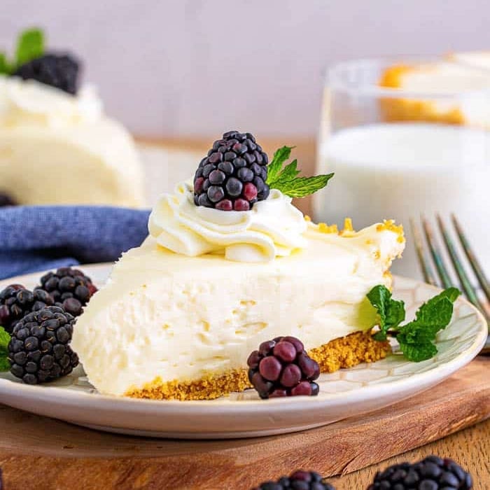 A slice of creamy cheesecake with a graham cracker crust, topped with whipped cream, a blackberry, and mint leaves-a perfect finish to your Easter Dinner. More blackberries and mint garnish the plate; milk and another cheesecake slice sit in the background.