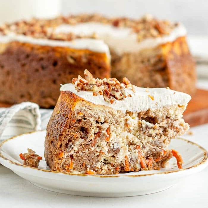 A slice of carrot cake topped with cream cheese frosting and chopped pecans sits on a plate, perfect for ending a festive Easter Dinner, with the rest of the cake in the background.