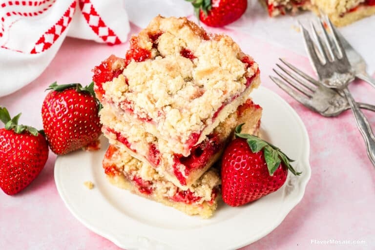 A stack of three strawberry crumb bars sits on a white plate, surrounded by fresh strawberries-perfect for an Easter Dinner dessert. Two vintage forks and a white napkin with a red pattern are in the background on a light pink surface.