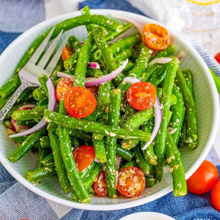 A bowl of fresh green bean salad with cherry tomatoes, red onion slices, and grated cheese-perfect for your Easter Dinner-served with a fork on a checkered cloth.