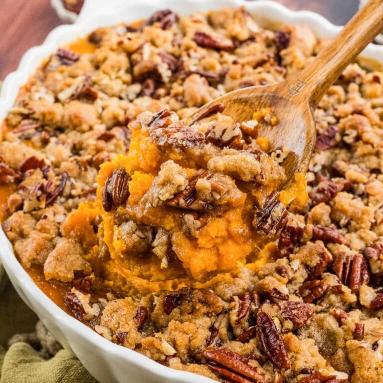 A close-up of a sweet potato casserole topped with a crumbly pecan streusel, perfect for Easter Dinner, with a wooden spoon lifting a serving from a white baking dish.