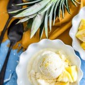 Overhead view of single serving of pineapple ice cream in white bowl with blue background with whole pineapple sliced vertically with orange napkin and 2 spoons.