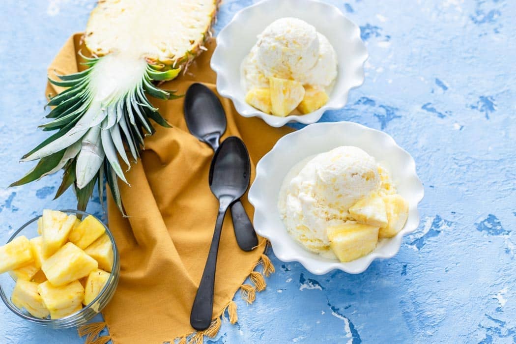 Two bowls of Pineapple Ice Cream with pineapple chunks, a bowl of sliced pineapple, two spoons, a yellow napkin, and a half-cut pineapple are arranged on a blue textured surface.