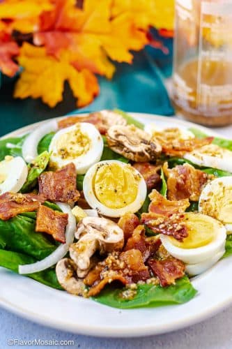 Spinach Salad with Warm Bacon Dressing on white plate with hard boiled eggs, bacon, sliced onions, and sliced mushrooms, with fall colored leaves on blue fabric and a bottle of bacon dressing in the background.