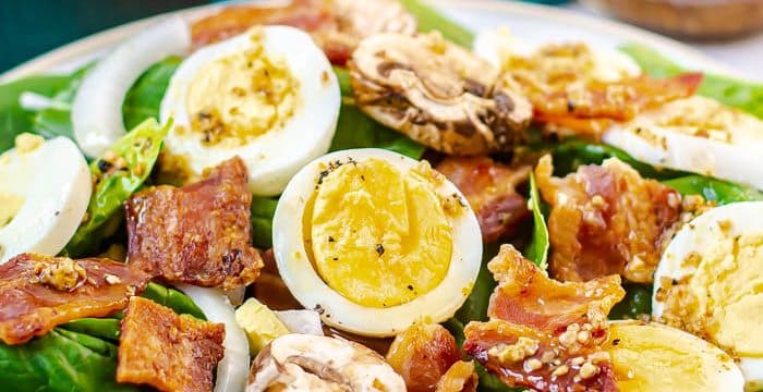 Spinach Salad with Warm Bacon Dressing on white plate with hard boiled eggs, bacon, sliced onions, and sliced mushrooms, with fall colored leaves on blue fabric and a bottle of bacon dressing in the background.