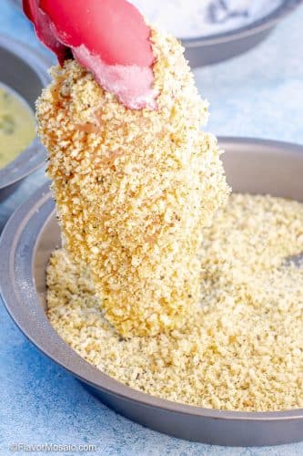 View of breaded chicken breast held in the air with red tongs over a pan of bread crumbs after the chicken breast has been covered with bread crumbs and before cooking.