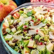 Glass bowl on dark gray background with apple and sliced pomegranate in the background and a tossed Shaved Brussels Sprouts Salad in bowl.