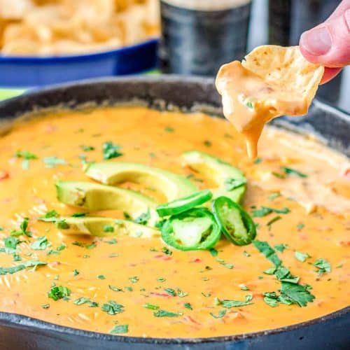 Skillet of loaded queso topped with sliced avocados,, jalapenos, and chopped cilantros, with a tortilla chip held over the skillet of queso with queso dripping off the chip into the skillet.