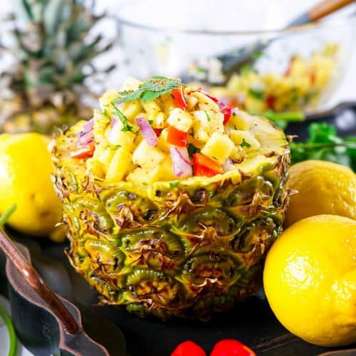 Side view of bottom half of pineapple, hollowed out and filled with pineapple salsa, surrounded by 3 whole lemons, 2 red chili peppers, cilantro, the top half of the pineapple in the back, along with a glass bowl of the remaining salsa.
