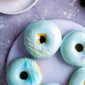Overhead view of a plate of donuts with a light blue glaze and yellow decorative frosting drizzled on top with small plate with single blue frosted donuts to the side in the upper left corner.