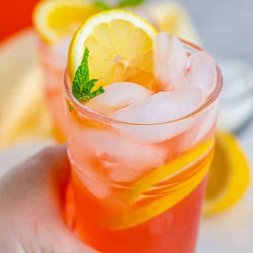 Someone's hand holding a glass of pink lemonade with ice cubes, sliced lemons, and a mint garnish.