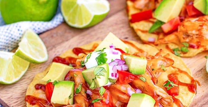 2 BBQ Chicken Tostadas on a cutting board topped with chicken, bbq sauce, chopped avocado and cheese. Surrounded by sliced limes, blue napkin whole avocado, whole lemon and whole tomatoes.