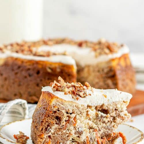 A single slice of Carrot Cake Cheesecake on a dessert plate with a view of the rest of the Carrot Cake Cheesecake and a glass bottle of milk in the background. Behind that is a white marble background.