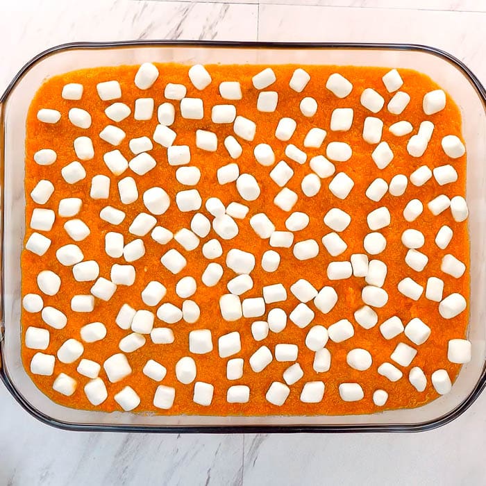 A glass baking dish filled with delicious Sweet Potato Casserole, topped with an even layer of mini marshmallows, sits on a white marble surface before it goes into the oven.