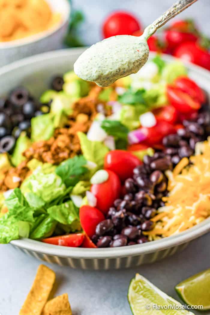 A spoon of cilantro lime dressing with a bowl of taco salad in which the ingredients are separated. Surrounded by tomatoes, limes and fritos.