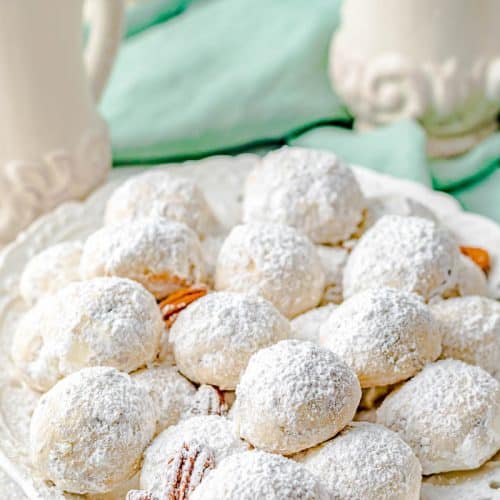 plate with about a dozen Mexican Wedding Cookies covered in powdered sugar and with a few pecan halves sprinkled on the plate, with 3 white vases and a teal napkin in the background.