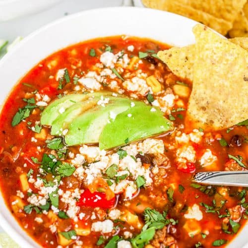 Overhead view of white bowl with Turkey Taco Soup with cheese crumbles, chopped cilantro, avocado slices and tortilla chips, and the bowl is surrounded by limes, tortilla chips, cilantro, and a partial view of a second bowl in the upper left corner.
