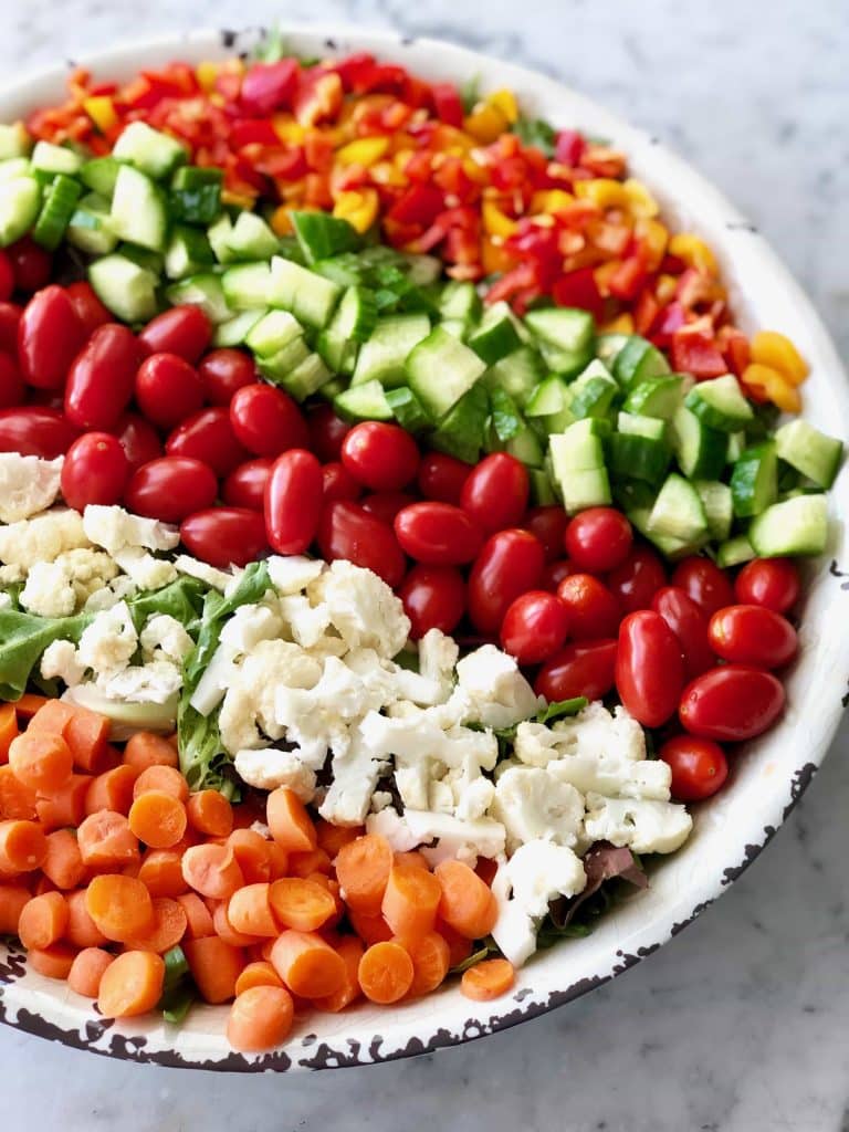Photo of a rainbow salad with the individual ingredients separated in rows on top of the salad in a rustic white bowl.