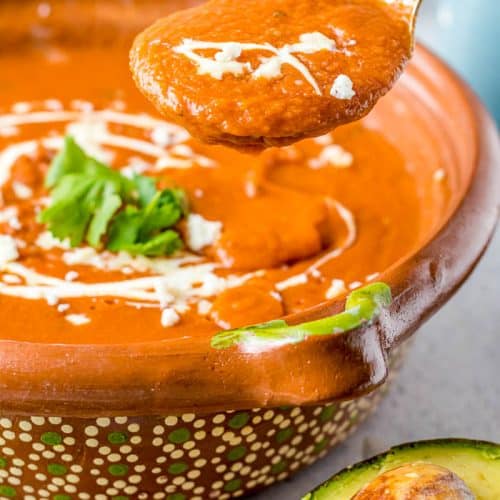 Spoonful of Sopa Tarasca - Pinto Bean Soup - with cream drizzled on top - held over a bowl of Sopa Tarasca drizzled with cream and garnished with cilantro. Half an avocado on the side.