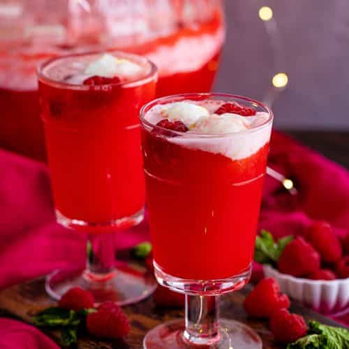 2 glasses of Christmas holiday punch with the punch bowl with more punch in the background.