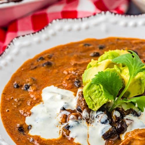 White bowl of Triple Pork Black Bean Soup topped with Mexican Cream, chopped avocado, and cilantro