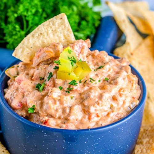 Blue bowl of Dill Pickle Cheeseburger Dip topped with chopped dill pickles surrounded by tortilla chips and green parsley in the background.