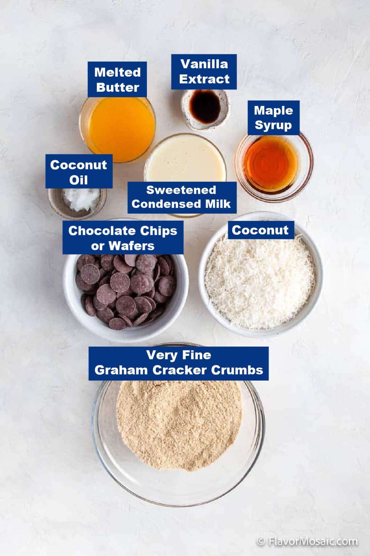 Top-down view of labeled baking ingredients in bowls for Chocolate Coconut Bars, including melted butter, vanilla extract, maple syrup, coconut oil, sweetened condensed milk, chocolate chips, shredded coconut, and fine graham cracker crumbs.