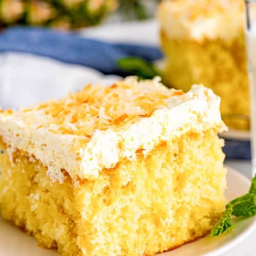 Close up view of 1 piece of Pina colada poke cake on a small white plate with a full glass of milk next to it with another piece of cake behind the milk and a blue and white napkin and a partial view of a pineapple in the background.