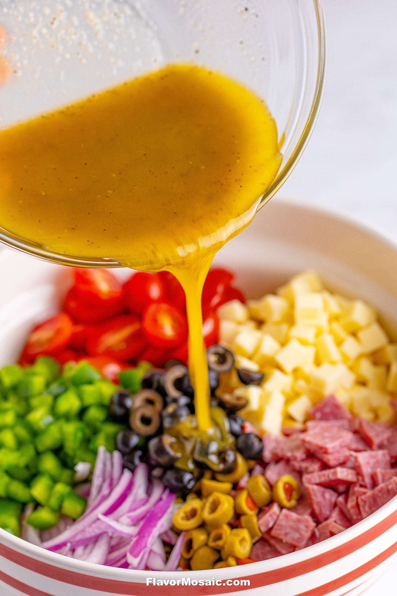 A bowl filled with chopped vegetables, cheese, olives, salami, and tri color rotini pasta, with a yellow vinaigrette being poured over the ingredients for a vibrant Tri Color Rotini Pasta Salad.