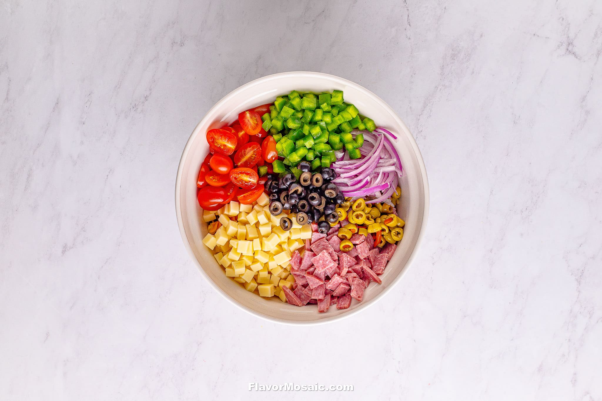 A white bowl on a marble surface filled with neatly arranged Tri Color Rotini Pasta Salad ingredients: cherry tomatoes, diced green bell pepper, sliced red onion, black olives, green olives, diced cheese, and sliced salami.