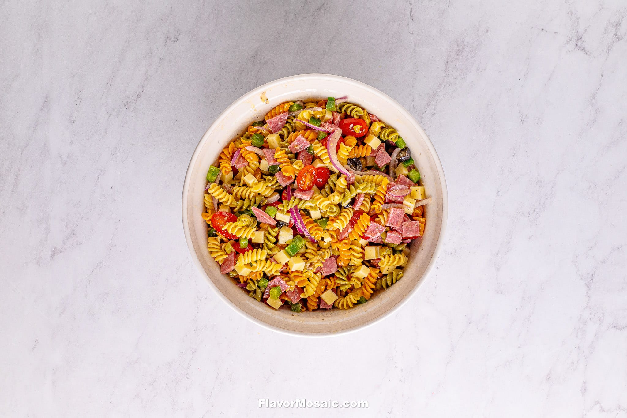 A bowl of Tri Color Rotini Pasta Salad filled with cherry tomatoes, green peas, cubed meat, and cheese sits on a light marble surface.