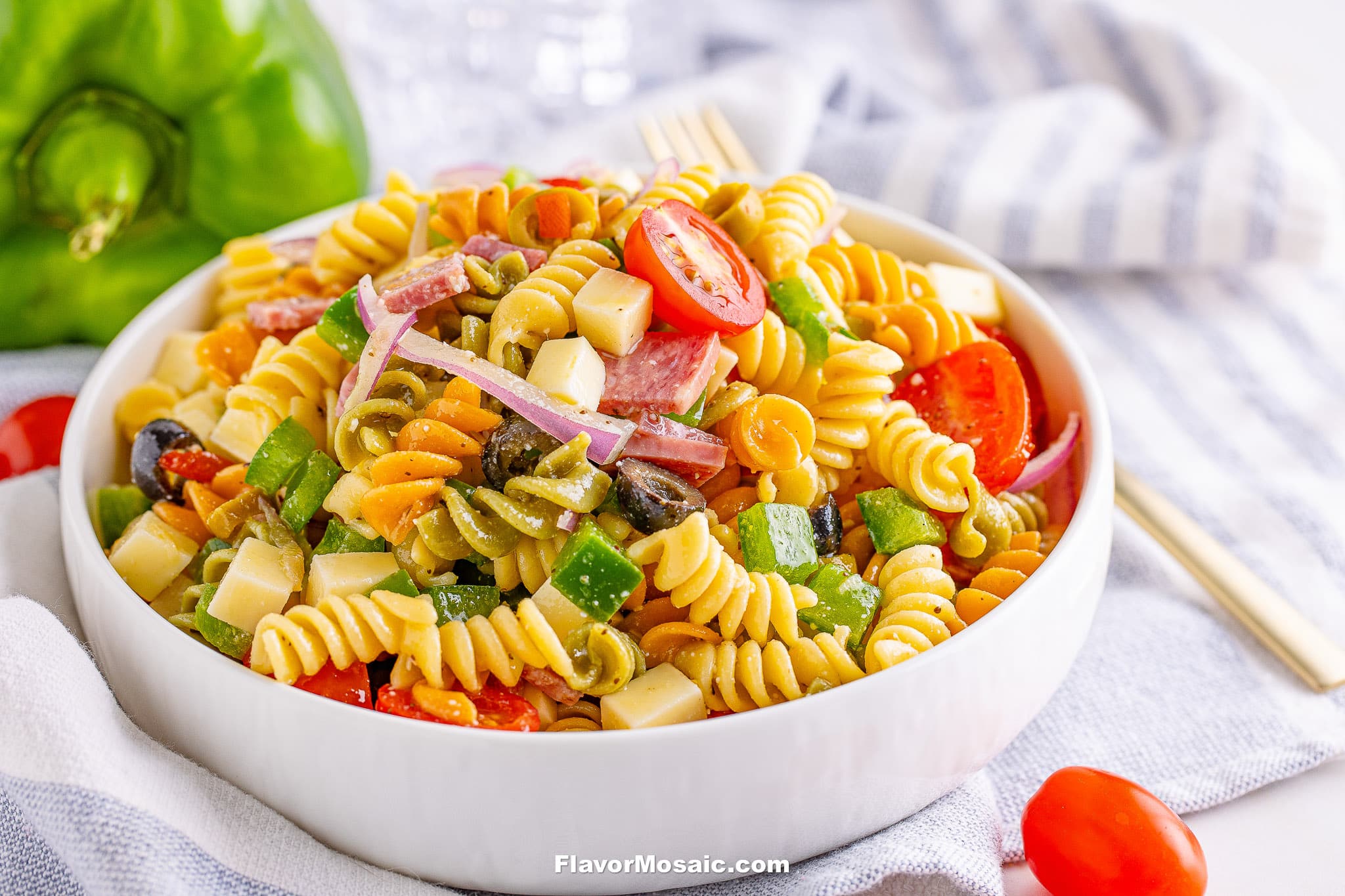 A bowl of Tri Color Rotini Pasta Salad with diced cheese, olives, sliced red onions, bell peppers, and cherry tomatoes is served on a white plate. A fork and napkin are in the background.