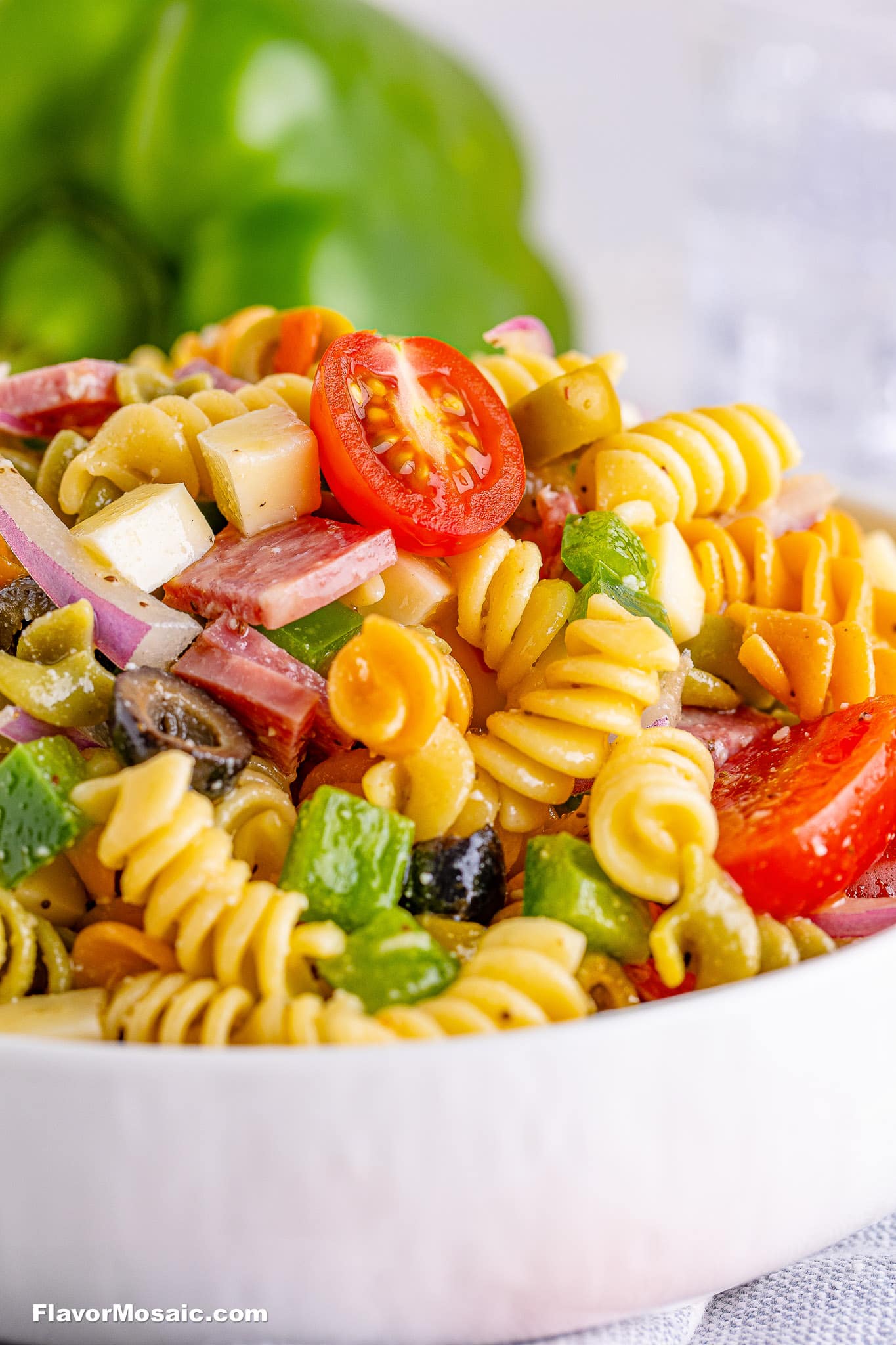 A close-up of a vibrant Tri Color Rotini Pasta Salad with cherry tomatoes, cubed cheese, sliced salami, red onion, green bell pepper, and black olives in a white bowl.