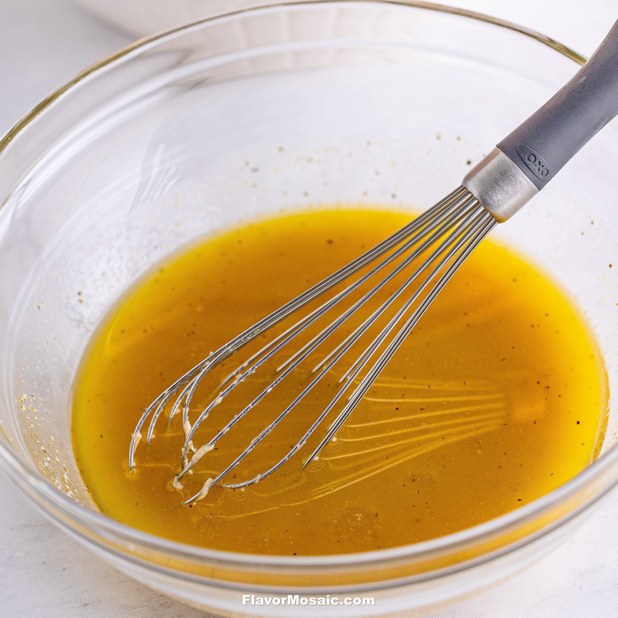 A metal whisk rests in a glass bowl filled with yellow vinaigrette dressing, perfect for tossing into a Tri Color Rotini Pasta Salad. The bowl sits on a white surface, and the vinaigrette is lightly speckled with seasonings.