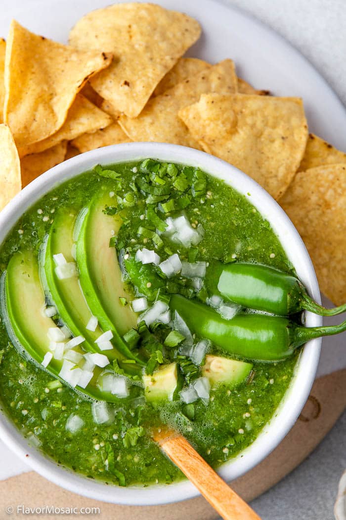 Overhead view of a bowl of salsa verde, green sauce, garnished with avocado slices, chopped onions, serrano peppers, and surrounded by tortilla chips.