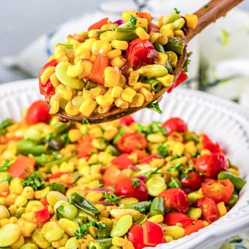 Side view of a white bowl with colorful succotash side dish, a mixture of corn, tomatoes, lima beans, and more. A spoonful of succotash is being lifted out of a large bowl of succotash.