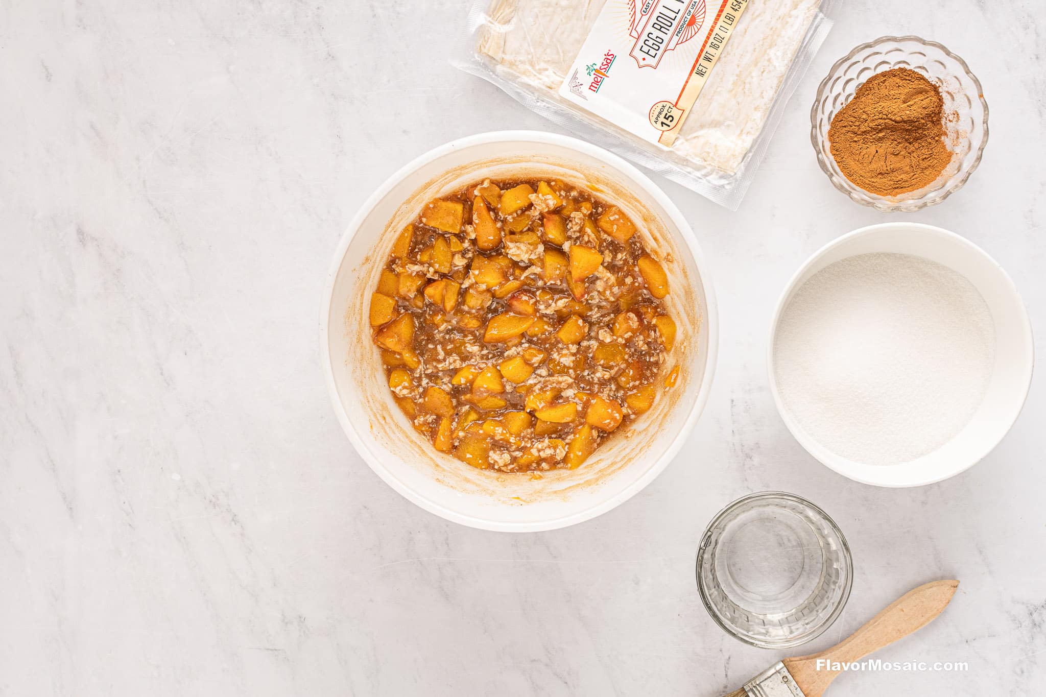 Top view of the ingredients for Peach Cobbler Egg Rolls: a bowl with peach filling, a packet of egg roll wrappers, a bowl of cinnamon, a bowl of sugar, a glass of water, and a pastry brush are all arranged on a light countertop.