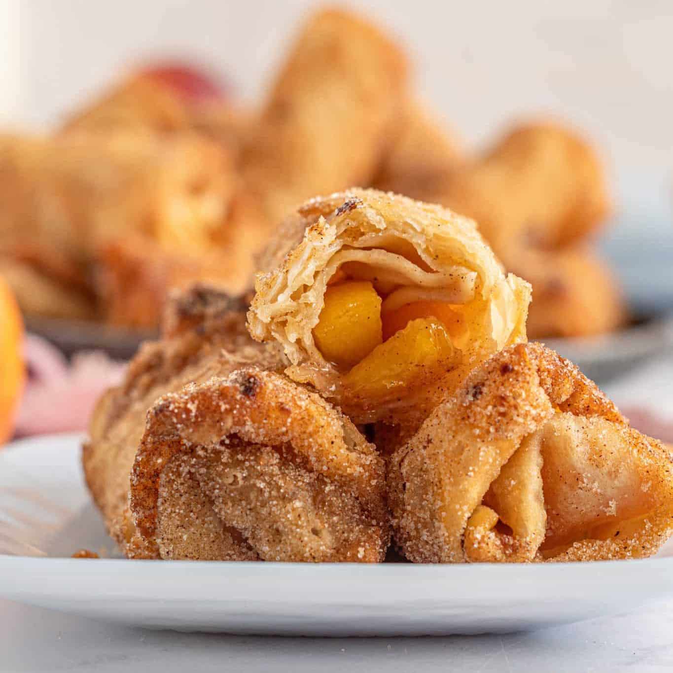 Close-up of golden, sugar-dusted Peach Cobbler Egg Rolls filled with juicy peach chunks, stacked on a white plate, with more pastries blurred in the background.
