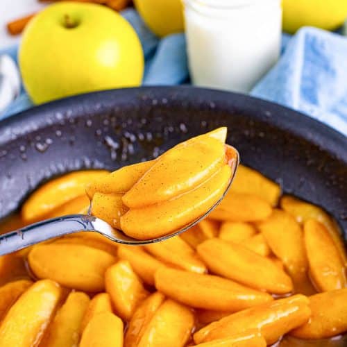 Fried apples in a skillet and a spoon holding a spoonful of fried apple slices above the skillet with 3 golden apples with a glass of milk and blue napkin in the background.