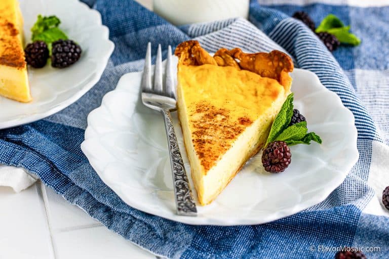 A slice of custard pie on a white plate, garnished with blackberries and a mint sprig, with a fork beside it. The plate rests on a blue plaid cloth.