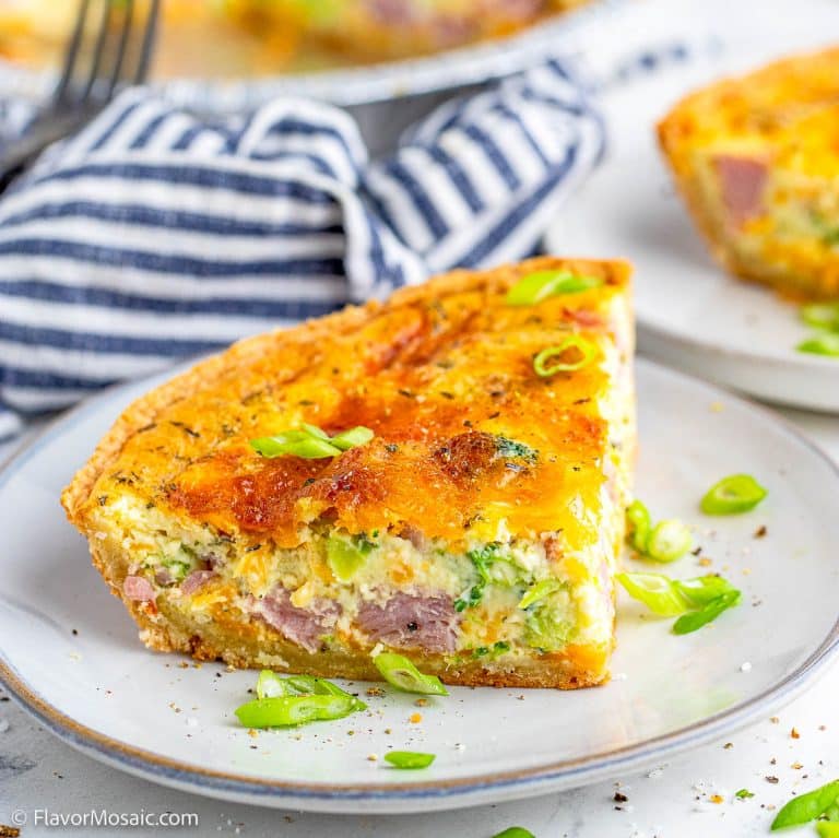 A slice of quiche with visible ham, cheese, and green onions sits on a plate. The quiche has a golden crust and is garnished with sliced green onions. A striped napkin is in the background.