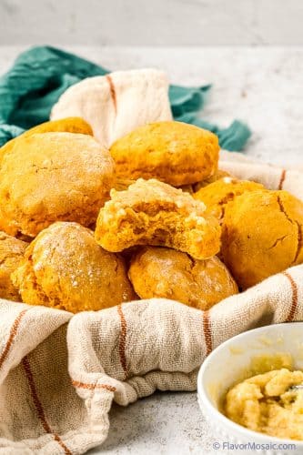 Side view of a towel covered bowl with at least half a dozen sweet potato biscuits with one biscuit sitting on top with a bite taken out of it and the top covered with honey butter. In the back is a green napkin.