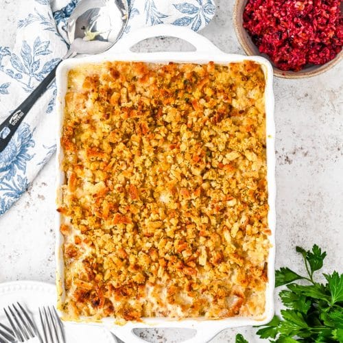 Overhead view of the entire chicken and stuffing casserole in a white casserole dish on a blue and white napkin on hte left on a white marble background, with cranberry sauce in hte upper left, parsleyin the bottom right, and 3 forks on a white plate in the bottom left.