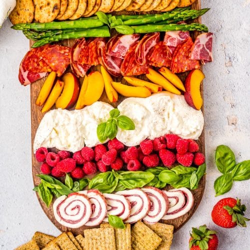Overhead view of a burrata board with burrata, raspberries, proscuitto, basil leaves, asparagus, crackers, and salami.
