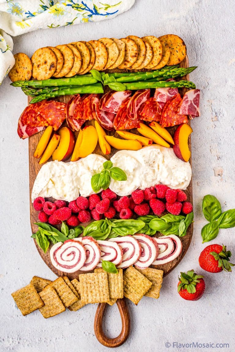 Overhead view of a burrata board with burrata, raspberries, proscuitto, basil leaves, asparagus, crackers, and salami.