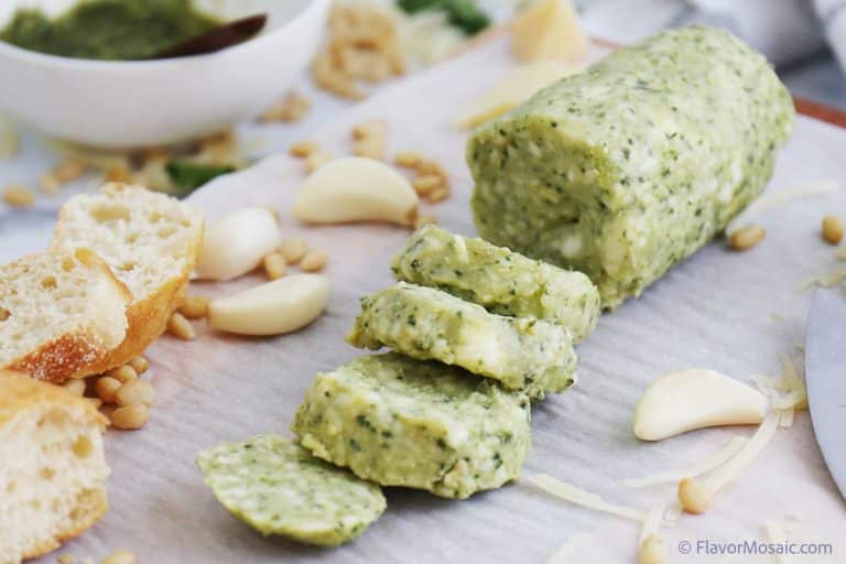 Pesto butter log cut into slices with garlic cloves, French baguette slices, and shredded parmesan cheese surrounding it.