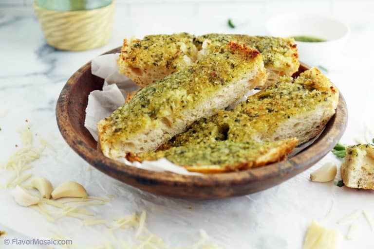 Pesto garlic bread slices on parchment paper in a wooden bowl sitting on top of white marble with garlic cloves and shredded parmesan surrounding it.