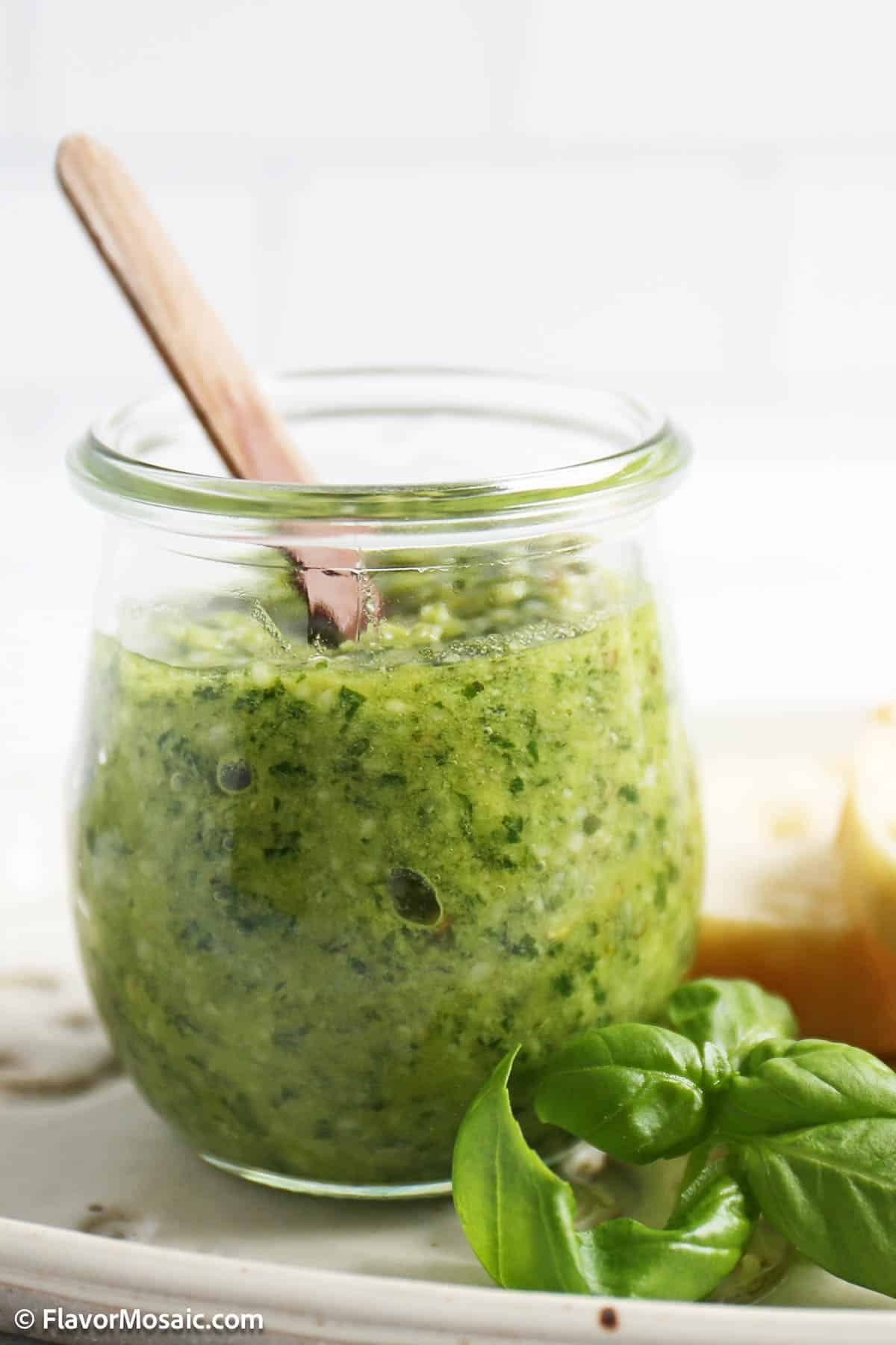 Side view of a clear glass jar of pesto with a wooden spoon in it. Next to the jar of pesto is fresh basil leaves and some slices of bread.